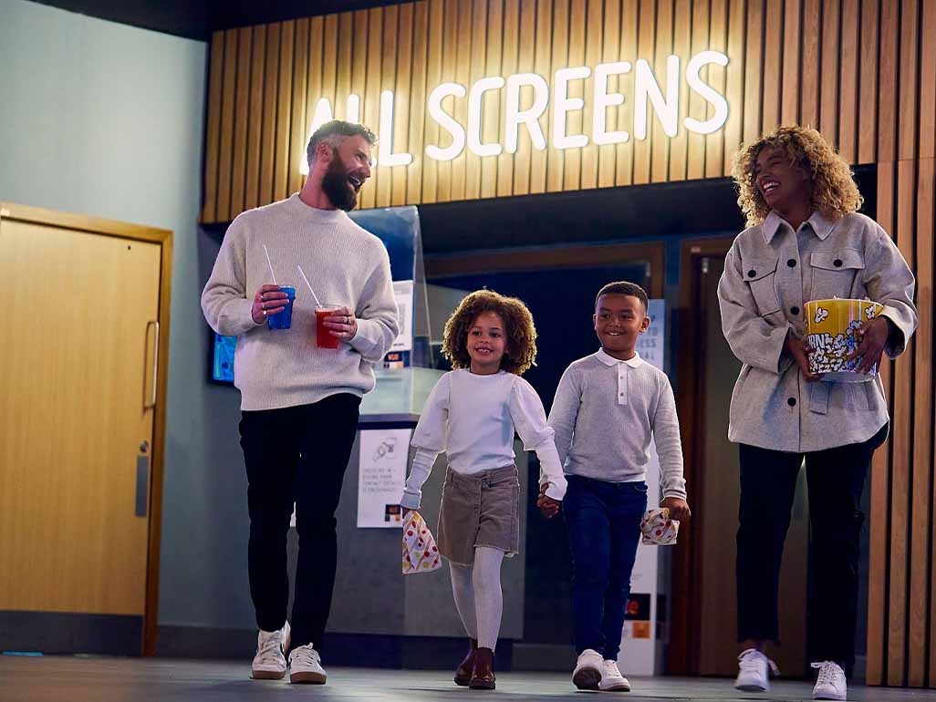A family of four walking out of the cinema screens laughing at each othe with drinks, popcorn and sweets